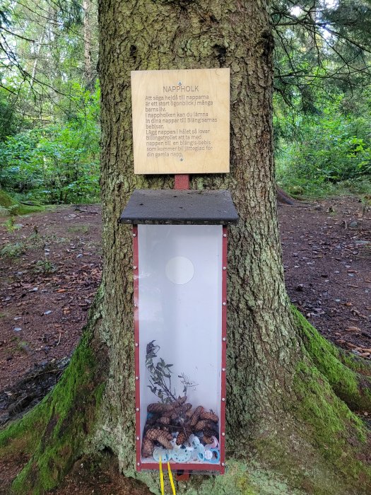 Vitrinskåp med nappar vid träd, skylt ovanför med text "NAPPHOLK", i en skogsmiljö.