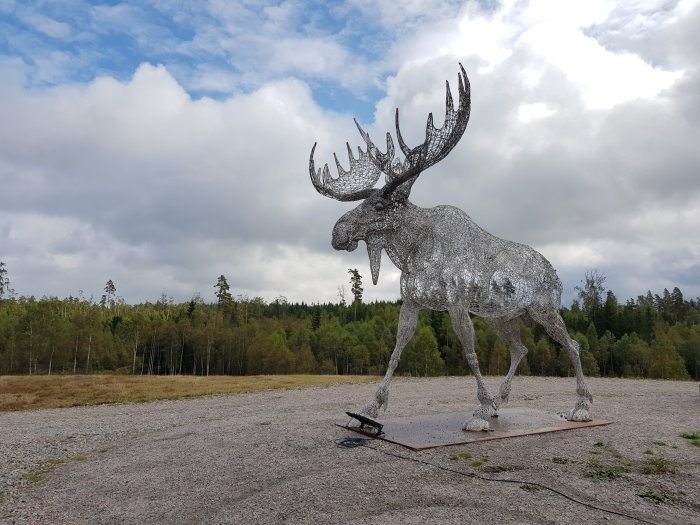 Stor trådskulptur av älg i naturlig miljö under molnig himmel.