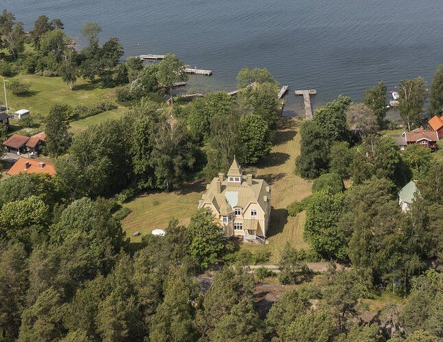 Flygbild av ett sekelskifteshus med sjötomt i Stockholms skärgård, omgivet av gröna träd och brygga.