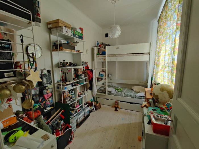 Stökigt compact living barnrum med en våningssäng, lådor, hyllor fulla av leksaker och böcker, och ett fönster med färgglada gardiner.