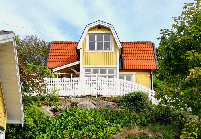 Gult hus med nybyggt vitt staket på en kulle omgiven av grönska.