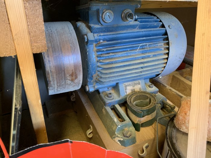 Blå industrimotor monterad på grönt stativ synlig bakom trästrukturer, i en verkstadsmiljö.