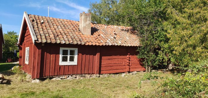 Ett rött 1700-tals uthus med slitet tegeltak och handslagna tegelpannor, omgivet av grönska.