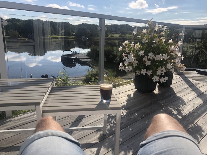 Altan med utsikt över en stilla sjö, kaffekopp på bordet och blommande planta, i soligt väder.