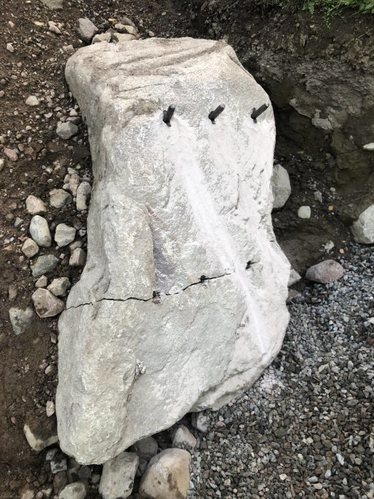 Stor sten med sprickor och insatta kilar redo för delning på en grusig mark.