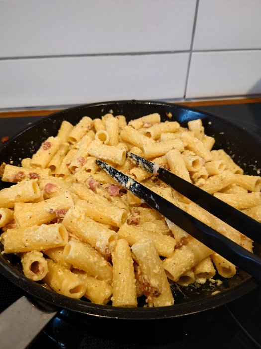 Grynig carbonara med pasta och bacon i en stekpanna, med matlagningsspadet.