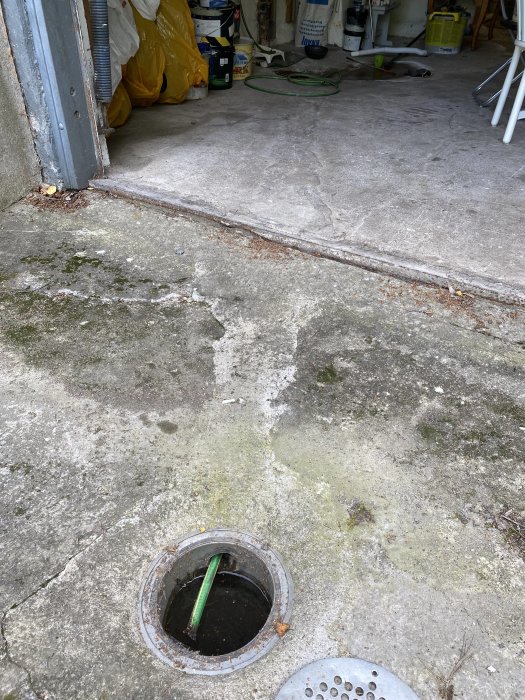 Avloppsbrunn med öppet lock på ett smutsigt garagegolv, nära garageporten, med en grön slang i brunnen.