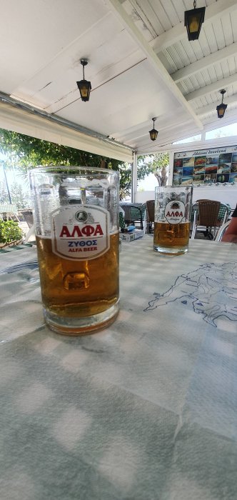 Två ölglas på ett bord med etiketter på främre glaset synligt märkt "ALFA BEER" utomhus på en veranda.