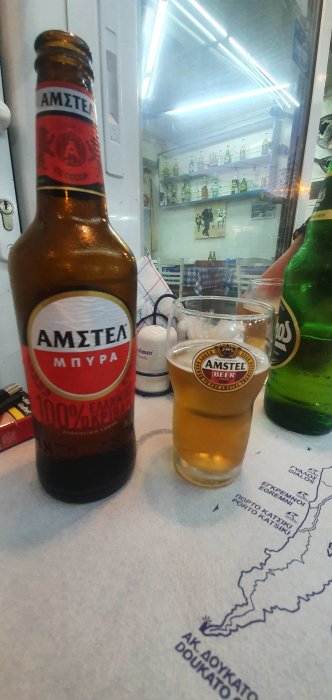 Flaska Amstel-öl och ett halvfullt glas framför en öppen kyl med flaskor, på en bordduk med kartmotiv.