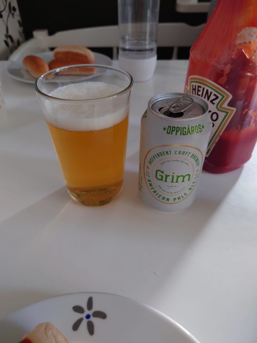 Ett glas öl bredvid en burk craftöl och en flaska ketchup på ett vitt bord med en korv i bakgrunden.
