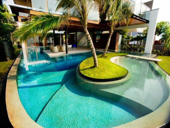 Lyxig pool som buktar runt en gräsö med ett träd, nära en elegant villa.