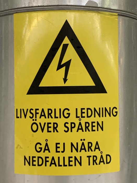 Varningsskylt med texten "LIVSFARLIG LEDNING ÖVER SPÅREN GÅ EJ NÄRA NEDFALLEN TRÅD" och en blixtsymbol.