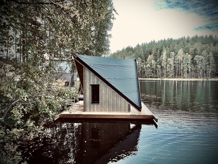 Färdigbyggt litet A-ramhus på en flotte vid en sjökant omgiven av skog.