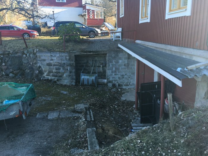 Garagenedfart med ojämn mark, mur och gammalt plåttak, bredvid ett rött traditionellt hus.