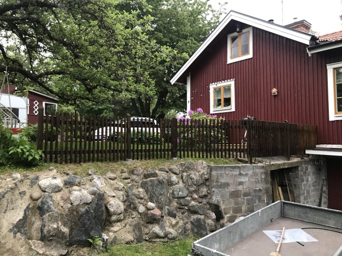 Äldre hus i rött med vita knutar, ojämn stenmur och trästaket, nedfart till garage under trädäck.