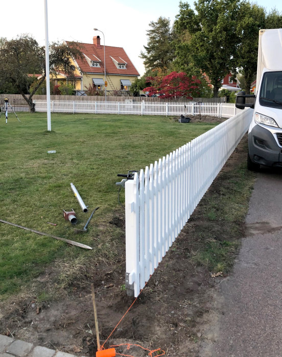 Nytt vitt staket längs gräsmatta med verktyg och byggmaterial synliga, hus i bakgrunden.