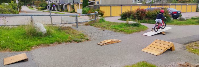 Barn cyklar på hemmagjorda träramper på en asfalterad väg i ett bostadsområde.