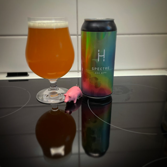 Ölglas med öl bredvid en burk märkt "SPECTRE DDH DIPA" och en liten rosa gris på en svart yta.