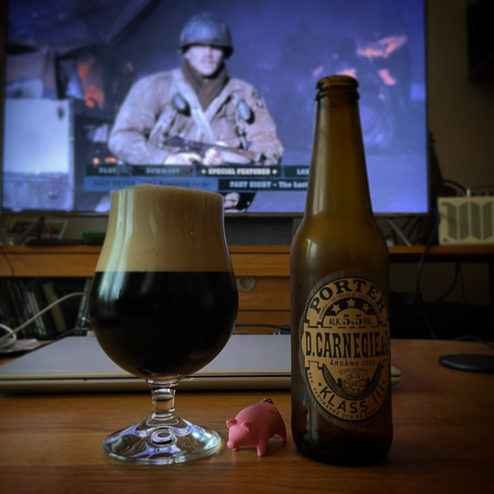 En porteröl i glas bredvid flaskan, med en gullig liten rosa spargris och en tv-skärm i bakgrunden.