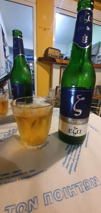 Två gröna ölflaskor av märket Eza och ett glas med gyllene dryck på ett bord med grekisk text.