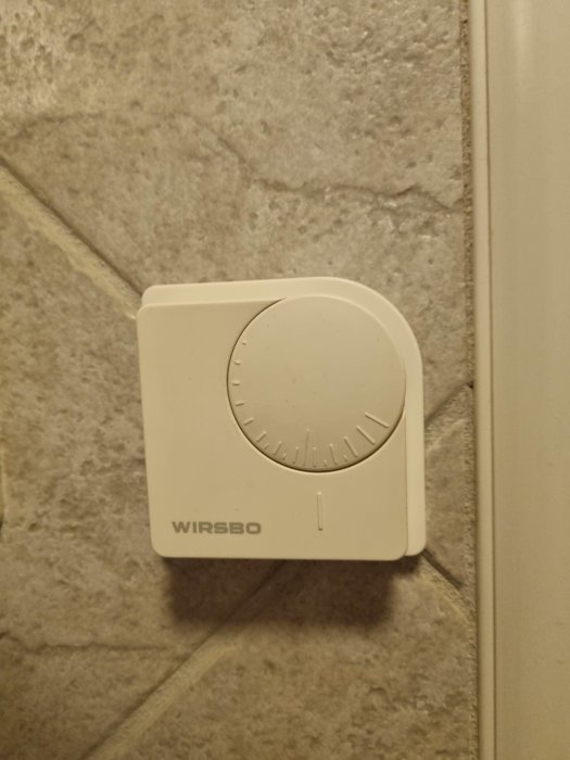 Vit Wirsbo-termostat på en vägg med gråa kakelplattor, ratten står på lägst värme och inget ljus syns.