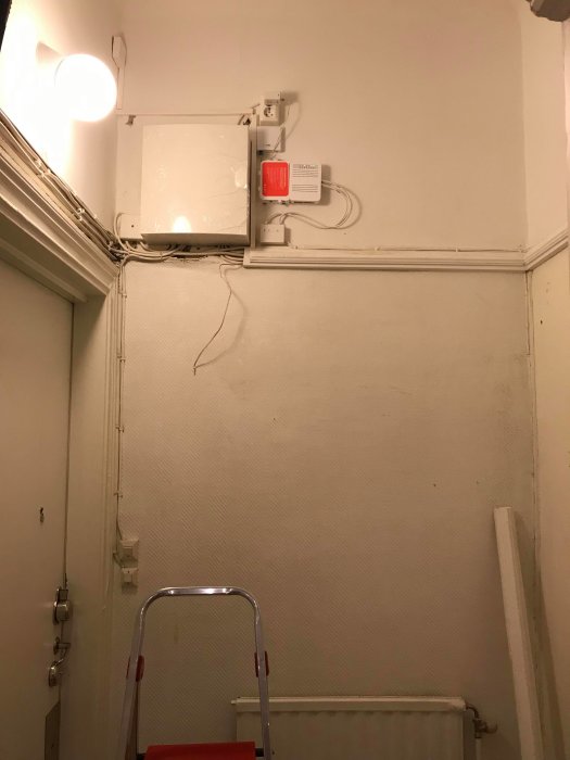 Hörn av en hall med avlägsnad vävtapet på väggarna, synliga ojämnheter och elinstallationer, intill en trappstege.