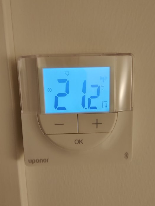 Uponor termostat med blå bakgrundsbelysning visar 21.2°C på displayen mot en beige vägg.