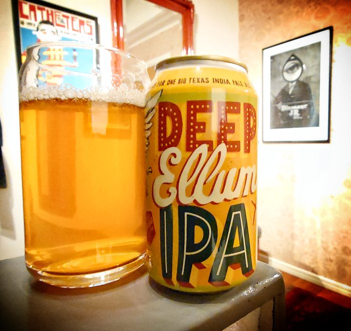 Ett glas öl bredvid Deep Ellum IPA-burk på bord, med suddig bild i bakgrunden.
