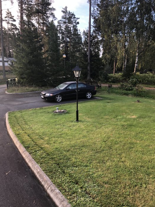 En utomhuslampa på en stolpe i en trädgård med en bil parkerad i bakgrunden och träd runtomkring.