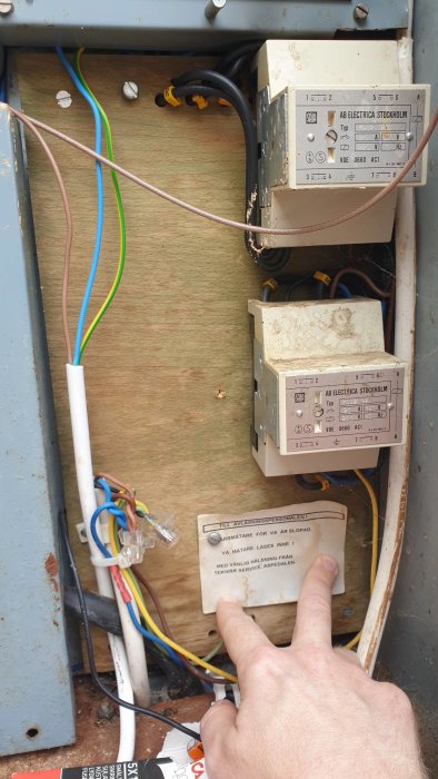 Elskåp med öppen dörr som visar flera elkomponenter och ledningar, hand pekar på etikett.