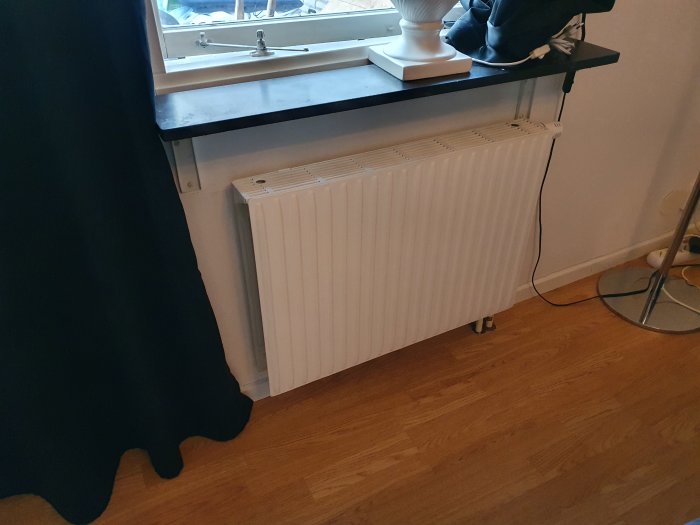 Värmeelement under fönster i ett rum med trägolv, gardin och skrivbord.
