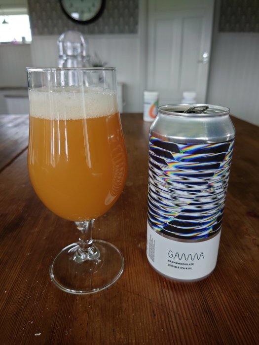 En ölglas fylld med grumlig, gul öl och en burk med konstnärlig design bredvid på ett träbord.