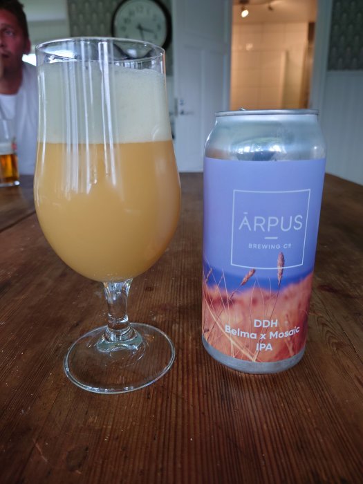 Ölglas med skummande öl bredvid en burk från Årpus Brewing Co, DDH Belma x Mosaic IPA, på träbord.