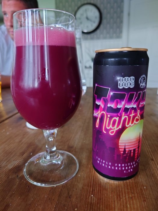 Ett glas med röd öl och en burk med etiketten "Tokyo Nights" på ett träbord, delvis synlig person i bakgrunden.