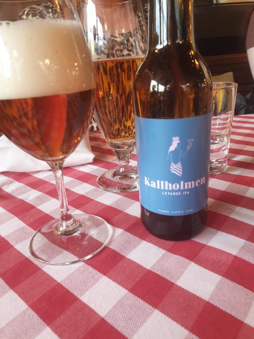 Flaska med Kallholmen Levande IPA på röd-vit rutig duk, omgiven av ölglas.
