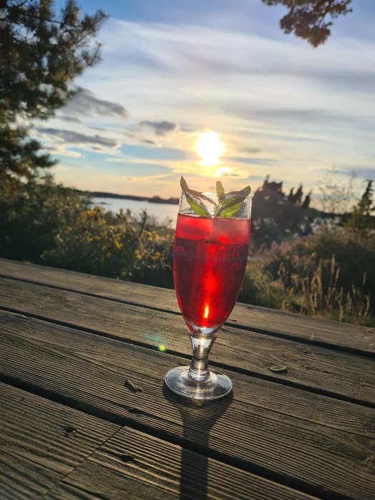 Ett glas med röd dryck och myntablad mot en solnedgångsbackdrop på en träyta.