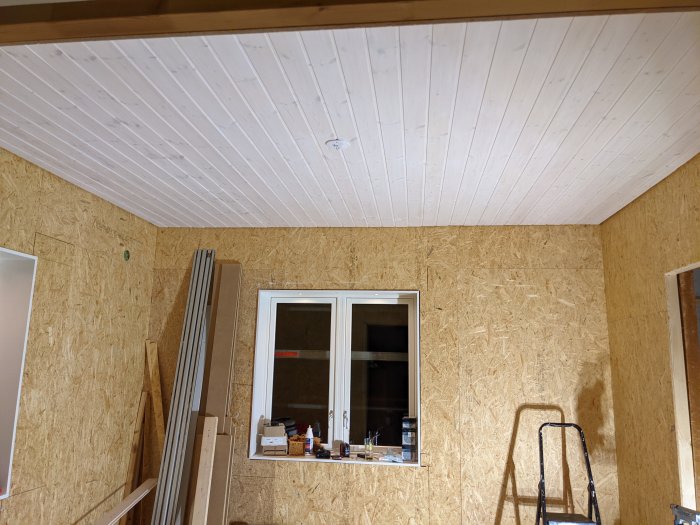 Nyinstallerad vit takpanel i ett rum med OSB-väggar, en fönstersektion och en stege vid sidan.