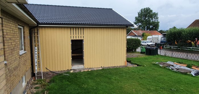 Bild på ett nyligen målat gult träskjul med öppet port vid ett hus och byggmaterial på marken.