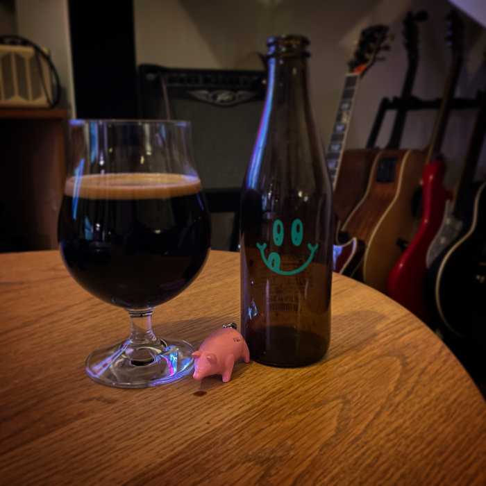 Ölstil med mörkt öl i snifterglas, tom ölflaska med leende motiv, småfigur av gris, gitarrer i bakgrunden.