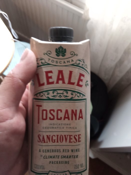 En hand håller en kartong av Toscanas Sangiovese rödvin nära ett fönster.