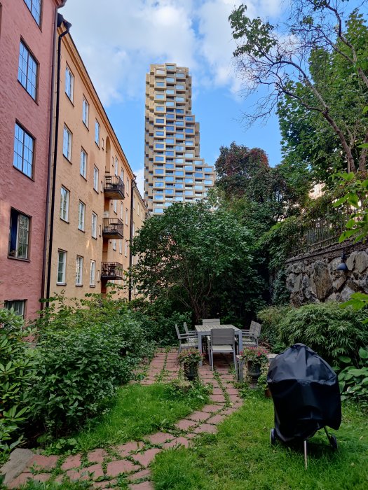 Trädgård med tegelstig framför äldre byggnader och modern höghus i bakgrunden under blå himmel.