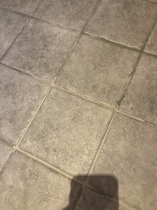 Närbild av kakelgolv med spruckna fogar och en lös platta, tecken på underliggande byggproblem.