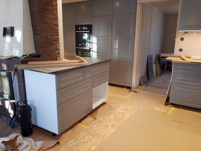 Pågående köksrenovering med installerade grå skåp och köksö, väntar på skåpsfronter, med burspråk.