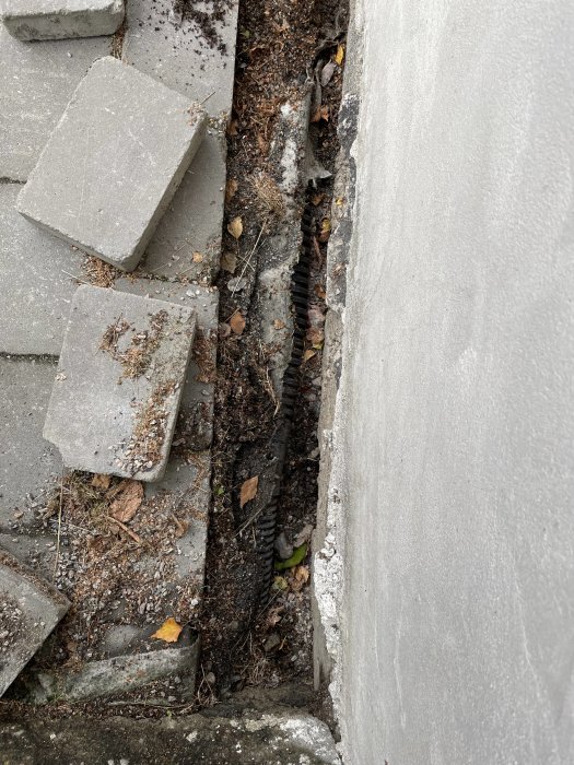 Skadade trädgårdsplattor vid husgrund med synligt dagvattenrör och hål intill.