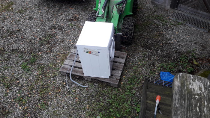 Vit tvättmaskin på pall vid en grön traktor utomhus, möjlig reservdelskälla.