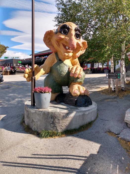 Färgglad stor figur av en trollkaraktär med käpp, utomhus intill en växt och stenfundament.