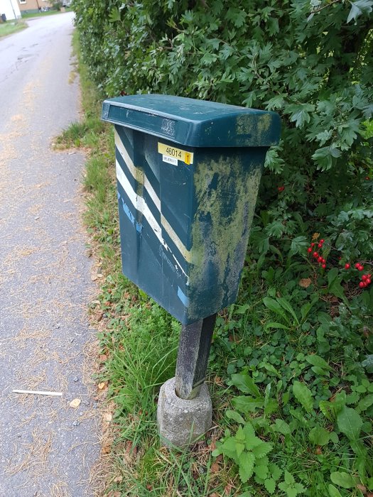 En gammal, möglig och rostig brevlåda vid en gata med grön vegetation i bakgrunden.