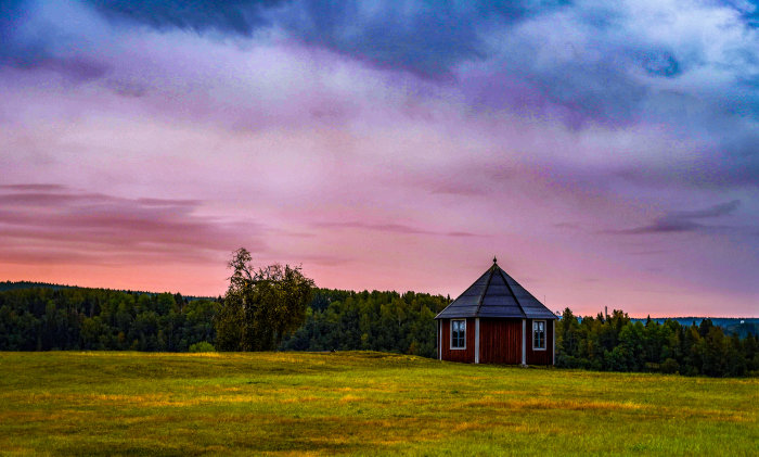 En liten röd stuga med svart tak på en öppen gräsmark under en dramatisk himmel vid skymning.