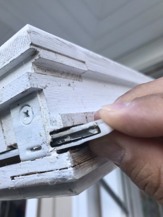 Hand håller en sliten plastdel på ett vitmålat fönster från EPA, skadad låsmekanism synlig.