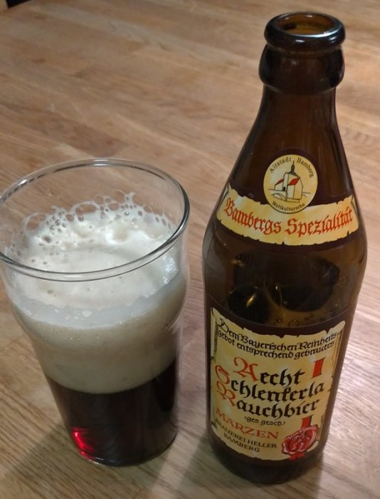 Flaska av Bamberg Spezialität Rauchbier bredvid ett glas med skummande mörkt öl på ett bord.